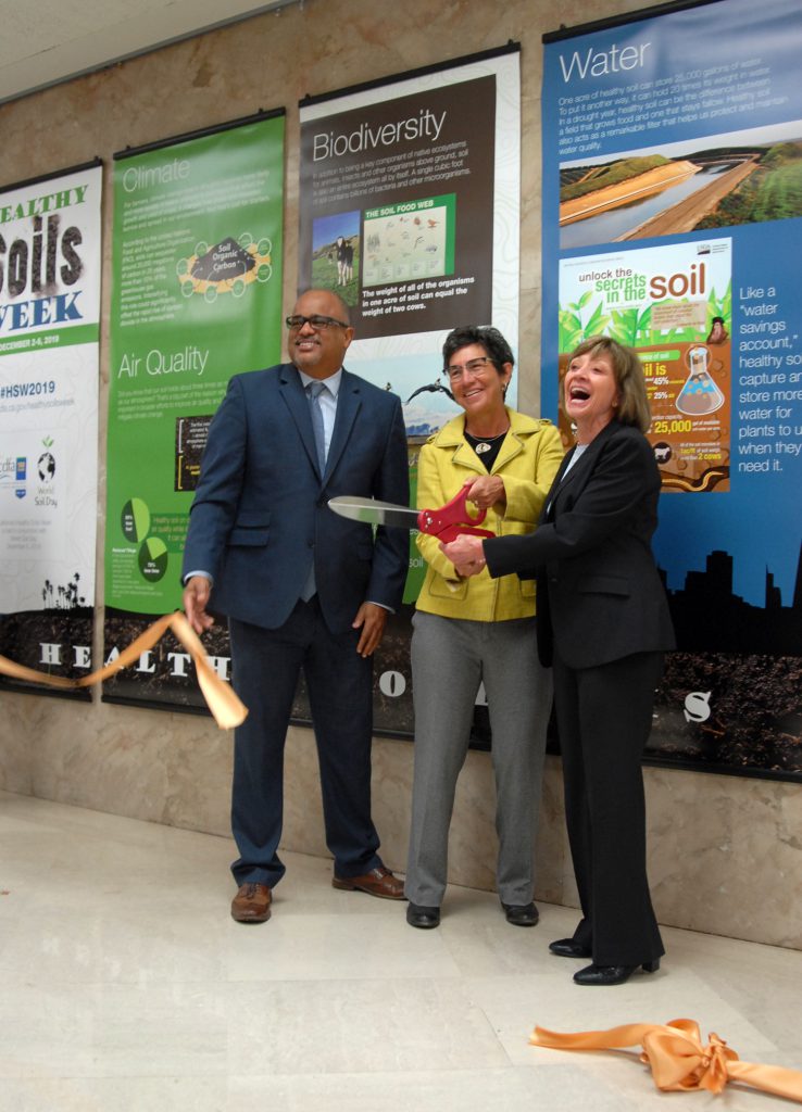 Photo of three officials cutting a gold ribbon in front of a set of seven banners hanging in a hallway at the State Capitol